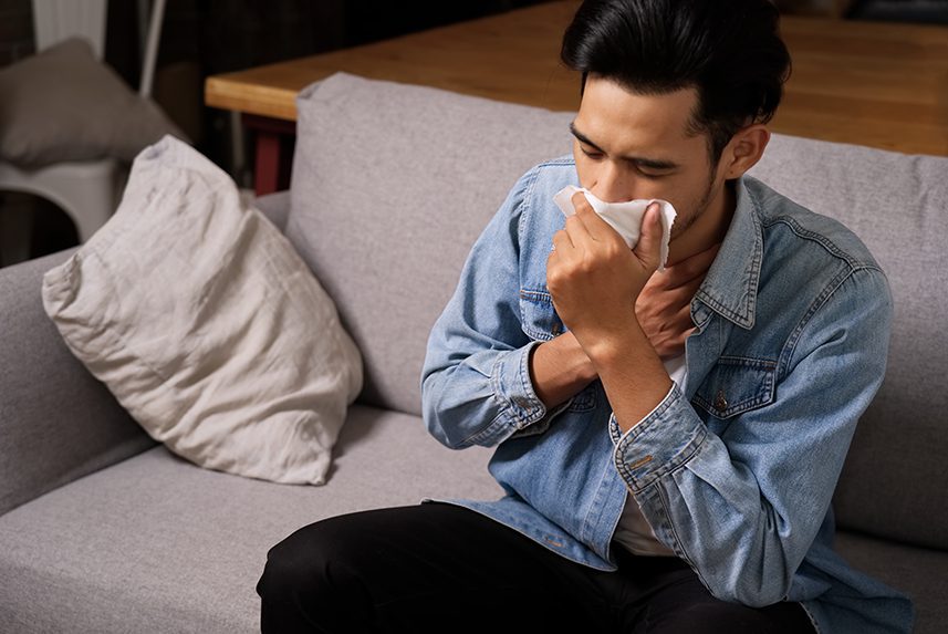 Person sitting on gray sofa while blowing their nose with a tissue.