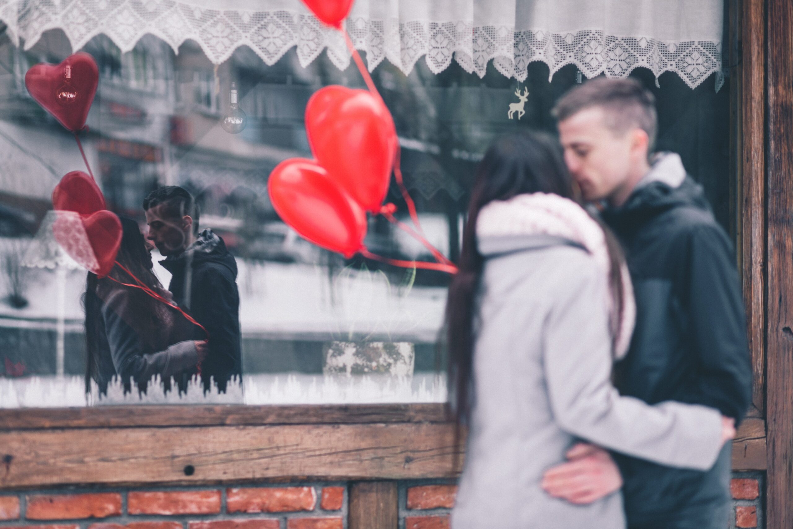 Photo of couple kissing with reflection on window pane. They are holding 3 heart shaped balloons.
