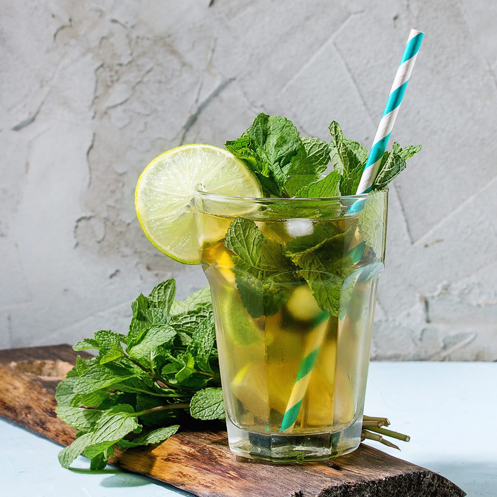 Glass of Iced green tea with lime, lemon, mint and ice cubes on wooden chopping board over light blue textured background.
