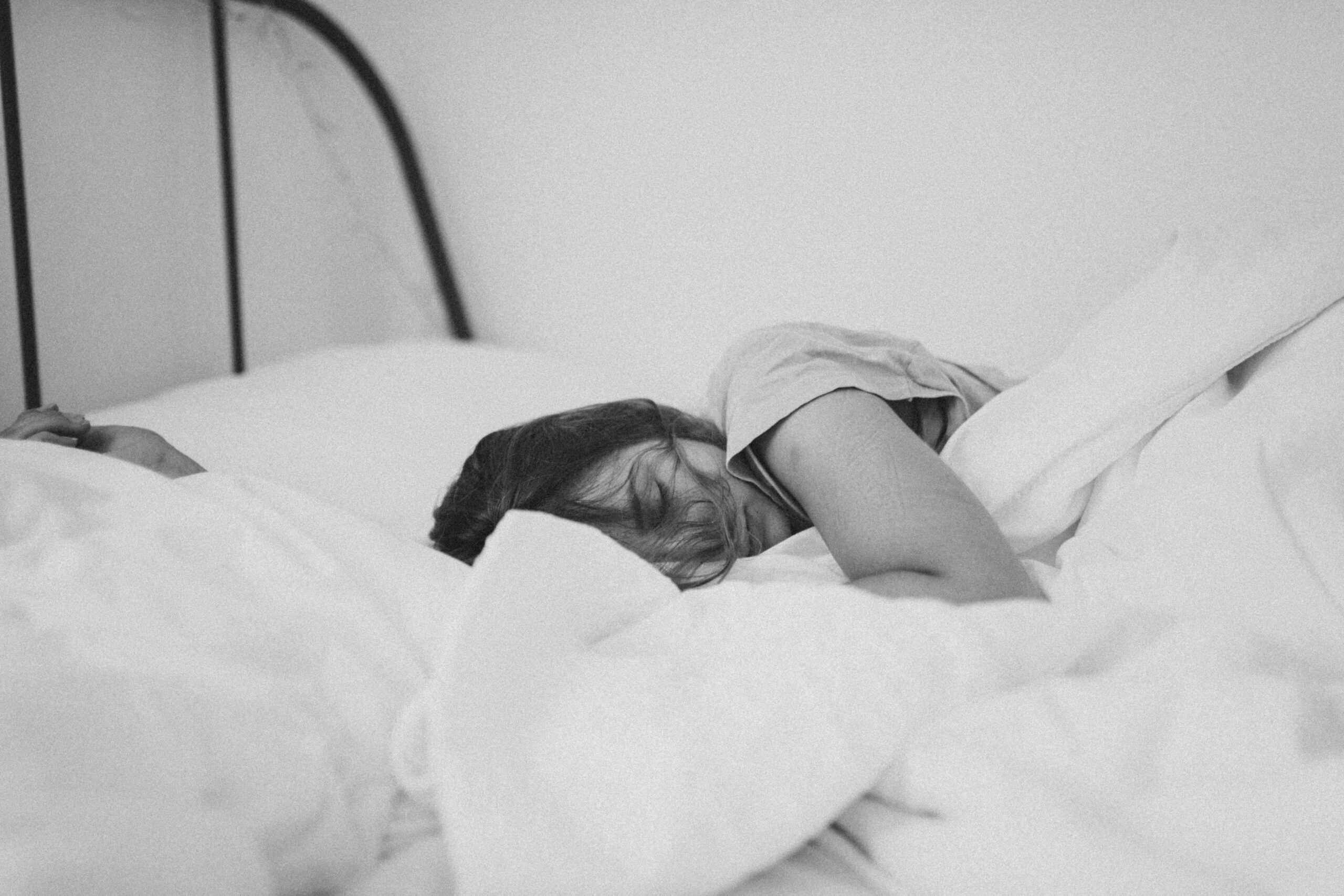 Black and white photo of person sleeping in bed. White sheets and pillowcases