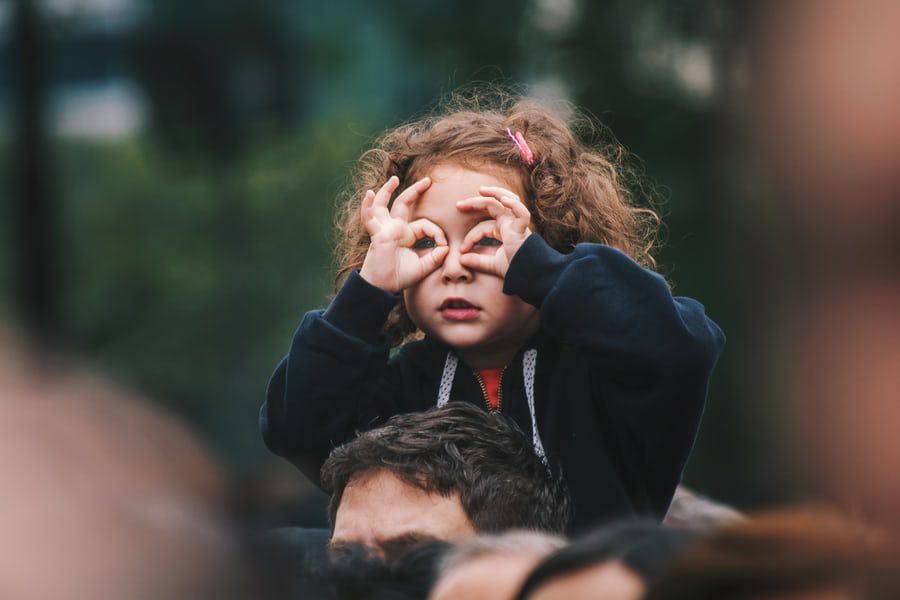 Young child sitting on parent's shoulders. Child made 2 circles with their thumb and pointer finger and placed them over their eyes like glasses