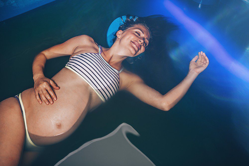 Pregnant woman floating in tank filled with dense salt water used in meditation, therapy, and alternative medicine.