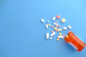 Medications and supplements spilling out of small amber vial on blue background.